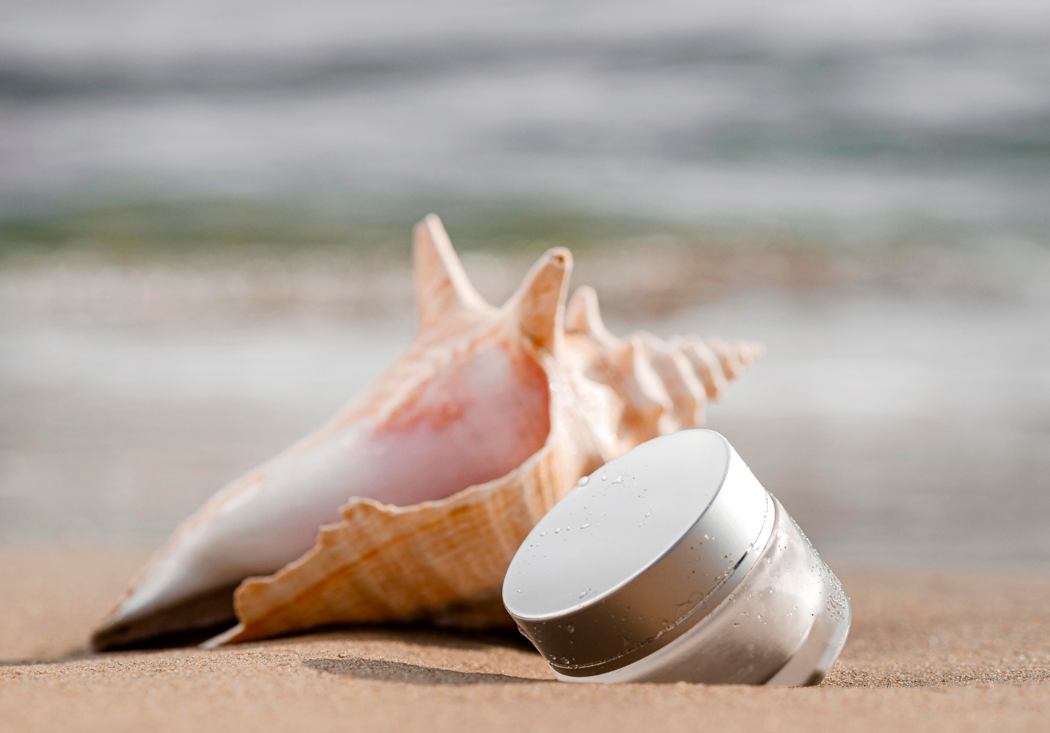 Une crème de soin de visage est posé à côté d'un coquillage sur une plage.